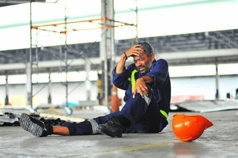 A worker in a blue jumpsuit sits on the floor of an industrial space, holding his head in pain. A fallen orange hard hat lies nearby.