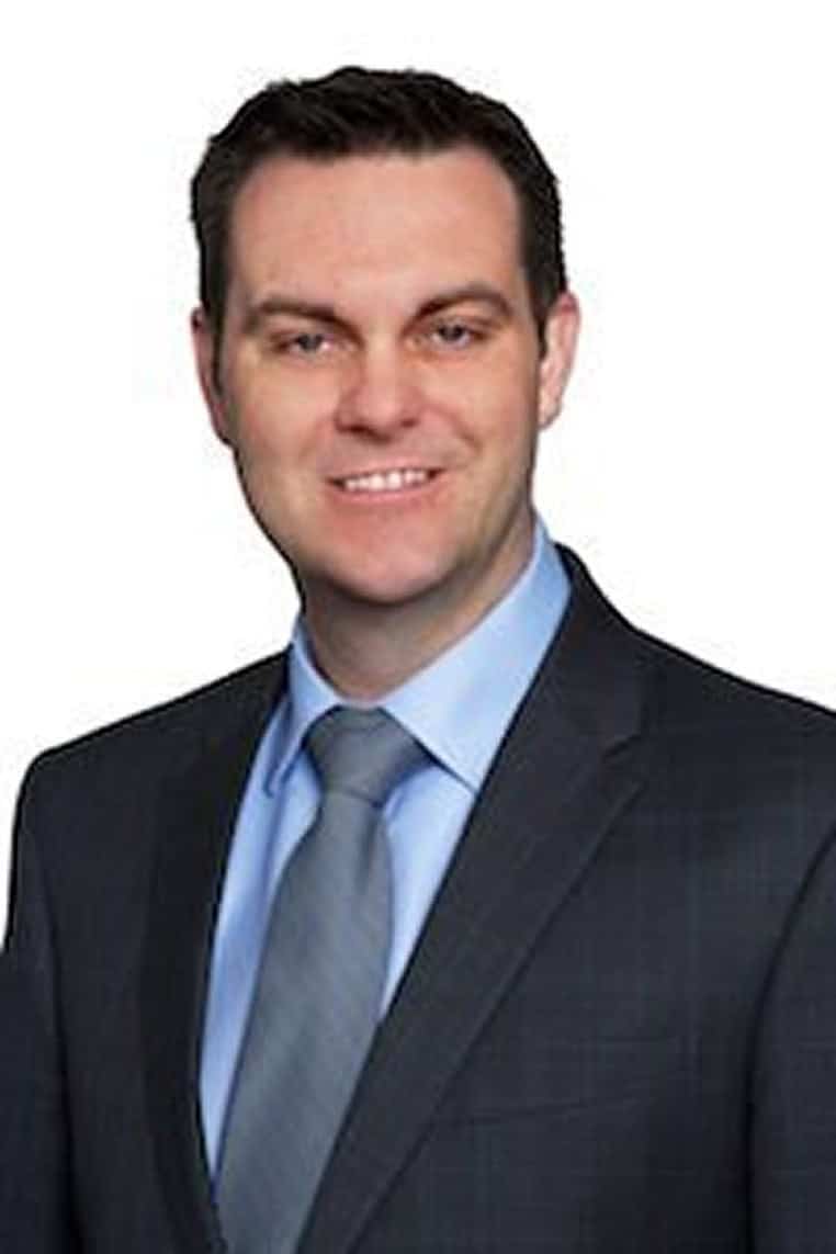 A man in a suit and tie smiling at the camera against a white background.
