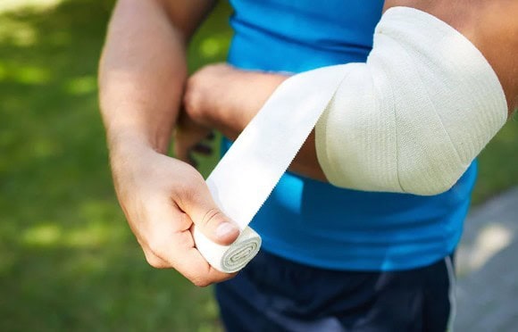 A person in a blue shirt wraps a bandage around their elbow outdoors.