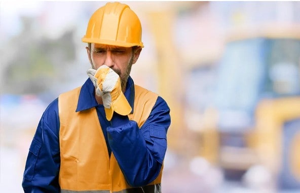 Construction worker in a hard hat and safety vest coughs into a gloved hand on a blurred construction site background.