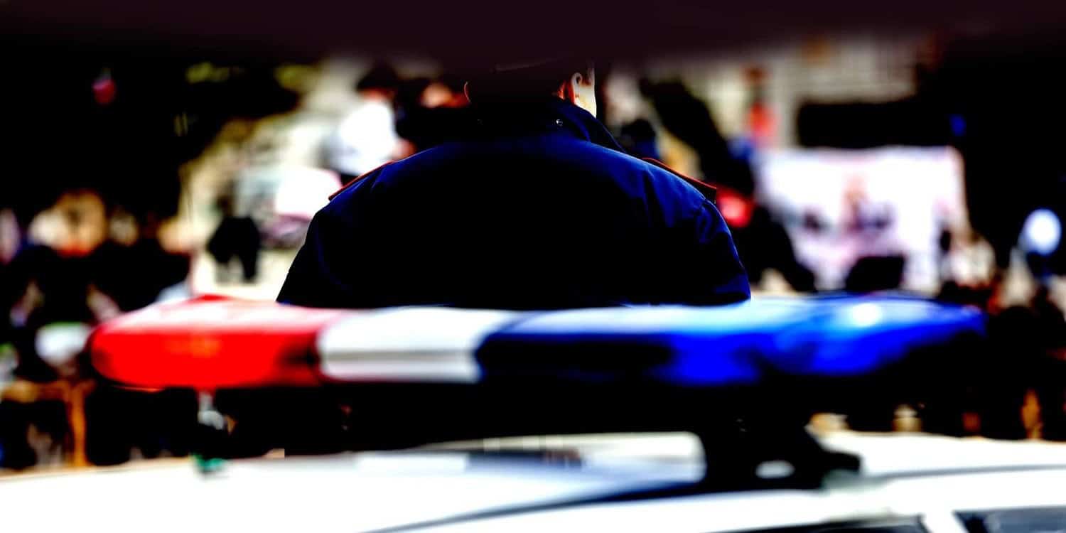 Police officer standing behind a patrol car with a red and blue flashing light bar.