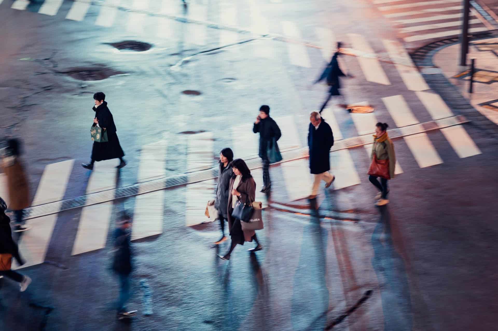 People walking across a city crosswalk at night, street lights illuminating the wet pavement.