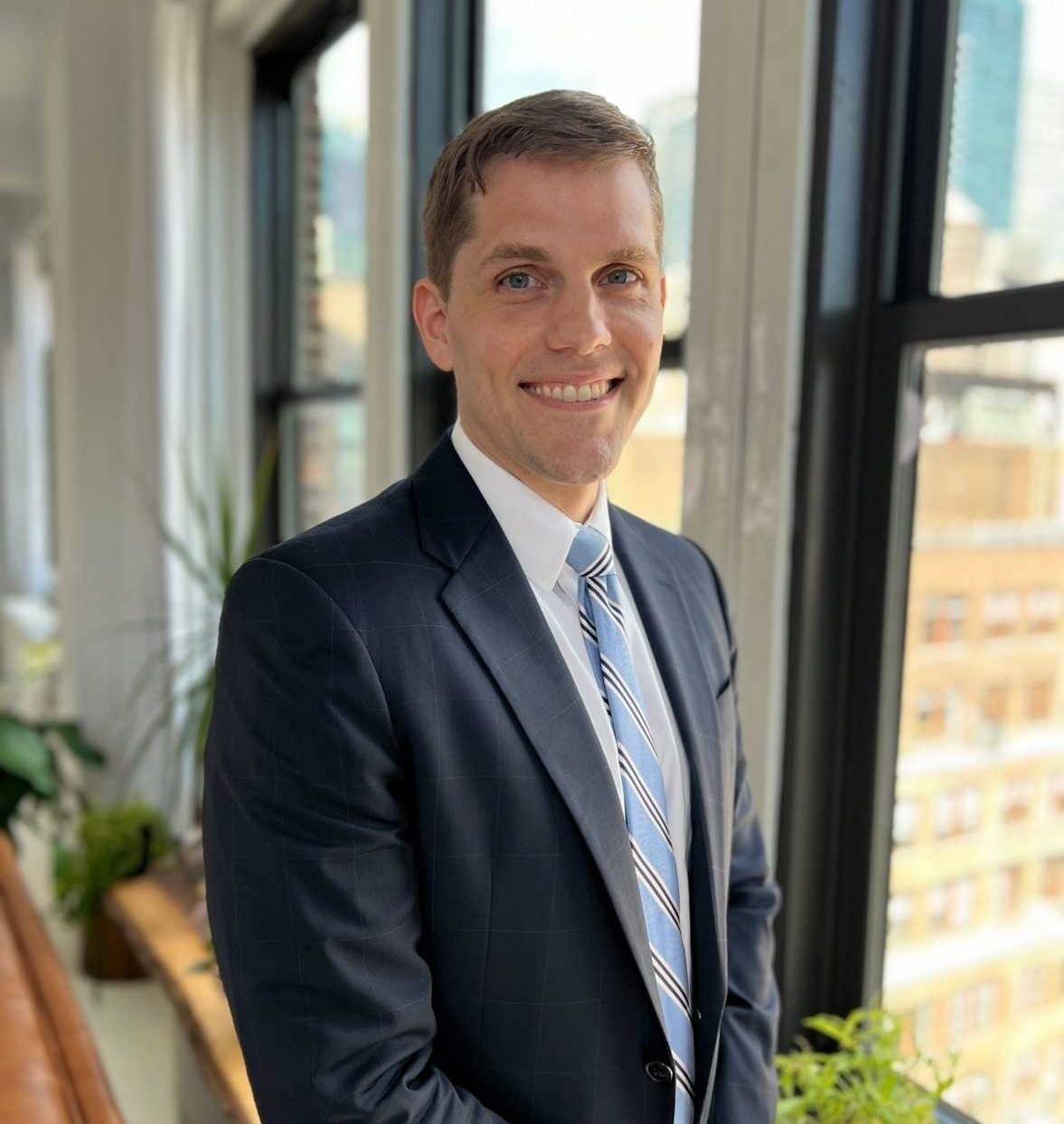 A man in a suit stands by a window in an office, smiling. There are plants on the windowsill and city buildings visible outside.